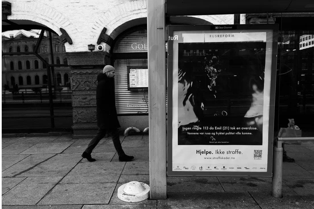 Passerby walking past a backlit Straff Skader bus shelter ad in a Norwegian city.