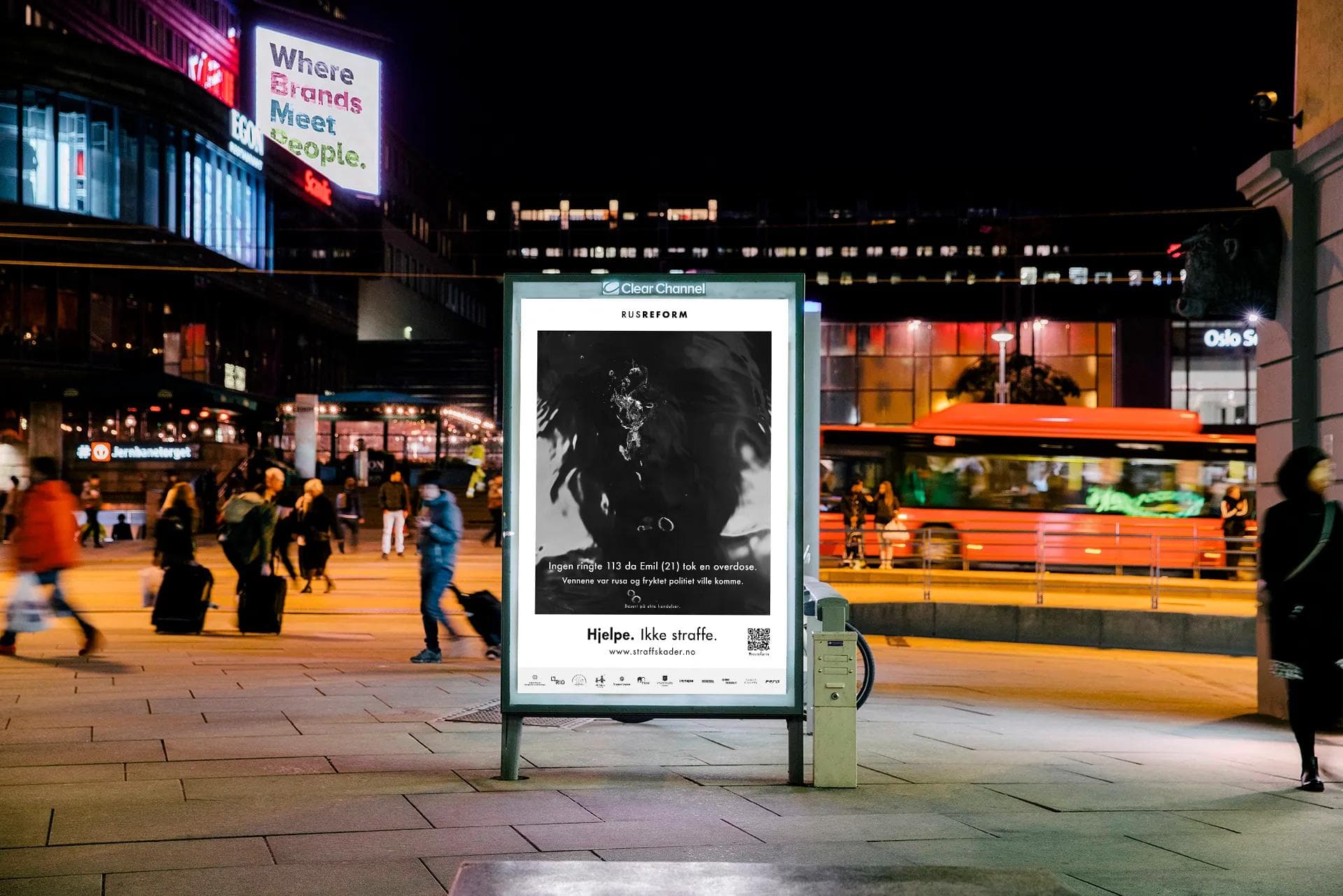 Nighttime at Jernbanetorget, Oslo — a lit campaign poster beside passing pedestrians and a red bus.